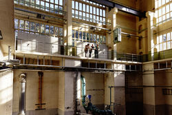 Auf dem Bild sind 3 Studierende in einer Industriehalle zu sehen wie sie an einem Geländer stehen und in die Halle hinunter blicken.