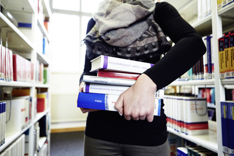 Auf dem Bild ist eine Studentin mit einem Stapel Büchern zwischen den Bücherregalen der Bibliothek zu sehen. 