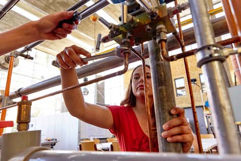 Studierende arbeiten in einer Maschinenhalle Auf dem Bild sieht man wie zwei Studierende an Rohren in einer Maschinenhalle arbeiten.