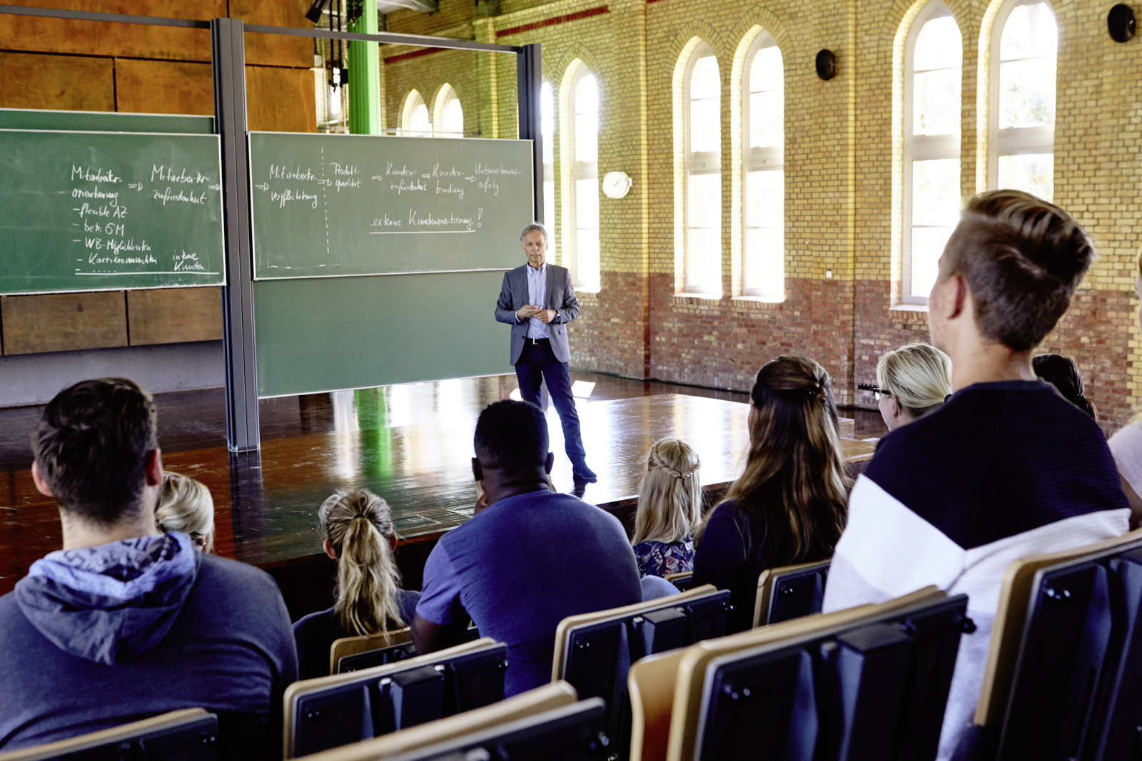 Auf dem Bild ist zu sehen wie ein Professor vor einer Tafel eine Vorlesung hält und die Studierenden ihm zuhören.